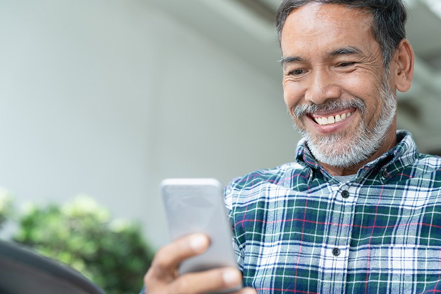 Older man with gray hair and beard smiles while looking at a smartphone, seated in a bright indoor setting.