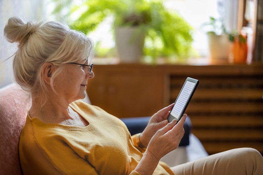 Older adult woman with gray hair and glasses sits comfortably on a couch, holding and reading from an e-reader in a softly lit living room with plants in the background.
