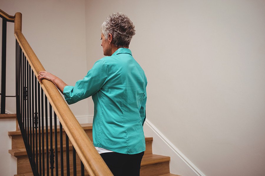 Older woman with short gray hair ascends a wooden staircase, holding the handrail for support in a well-lit home interior.