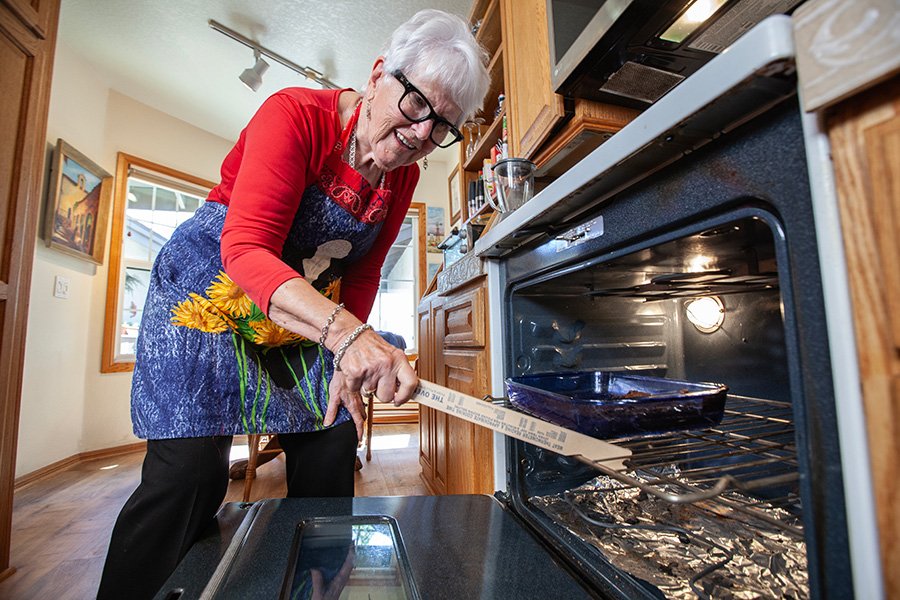 A woman in a blue apron opens a stove with a dish inside.