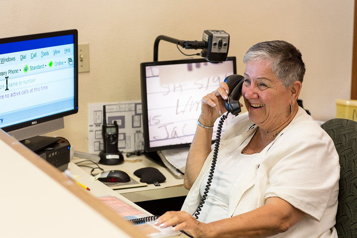 woman_on_phone A woman in a casual white shirt sits at a desk while answering a phone and smiling.